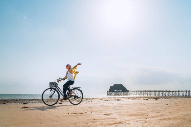 Young female dressed light summer clothes joyfully threw up her hand riding old vintage bicycle with front basket on the low tide ocean white sand coast on Kiwengwa beach on Zanzibar island, Tanzania.